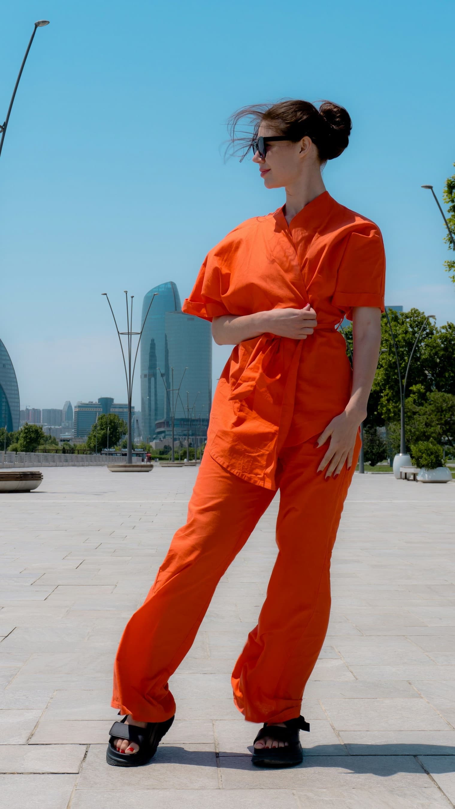 A woman in a bright orange wrap top and wide trousers, wearing dark sunglasses, stands in a plaza with glass towers, trees, and blue sky.
