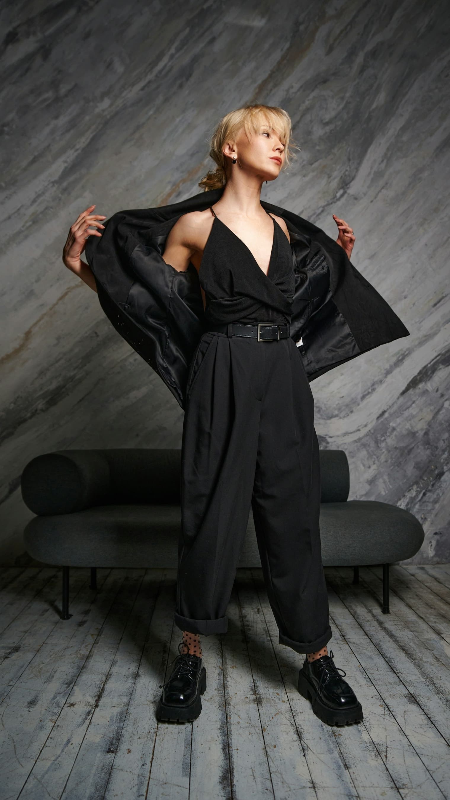 A woman in a black sleeveless wide-leg jumpsuit stands in a grey studio, a black blazer over her shoulders.