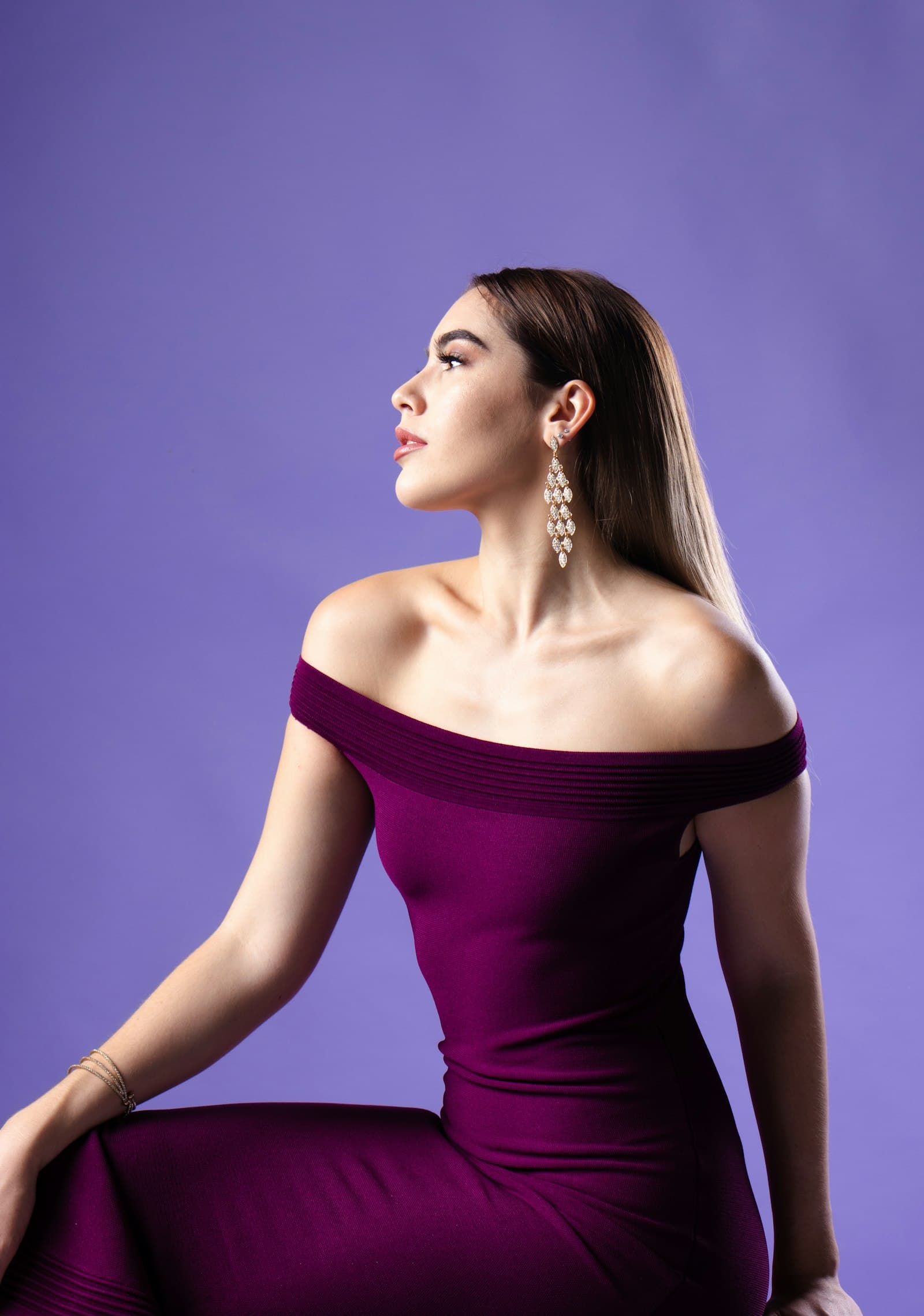 A woman in a fitted deep-purple off-the-shoulder dress poses against a solid purple backdrop.
