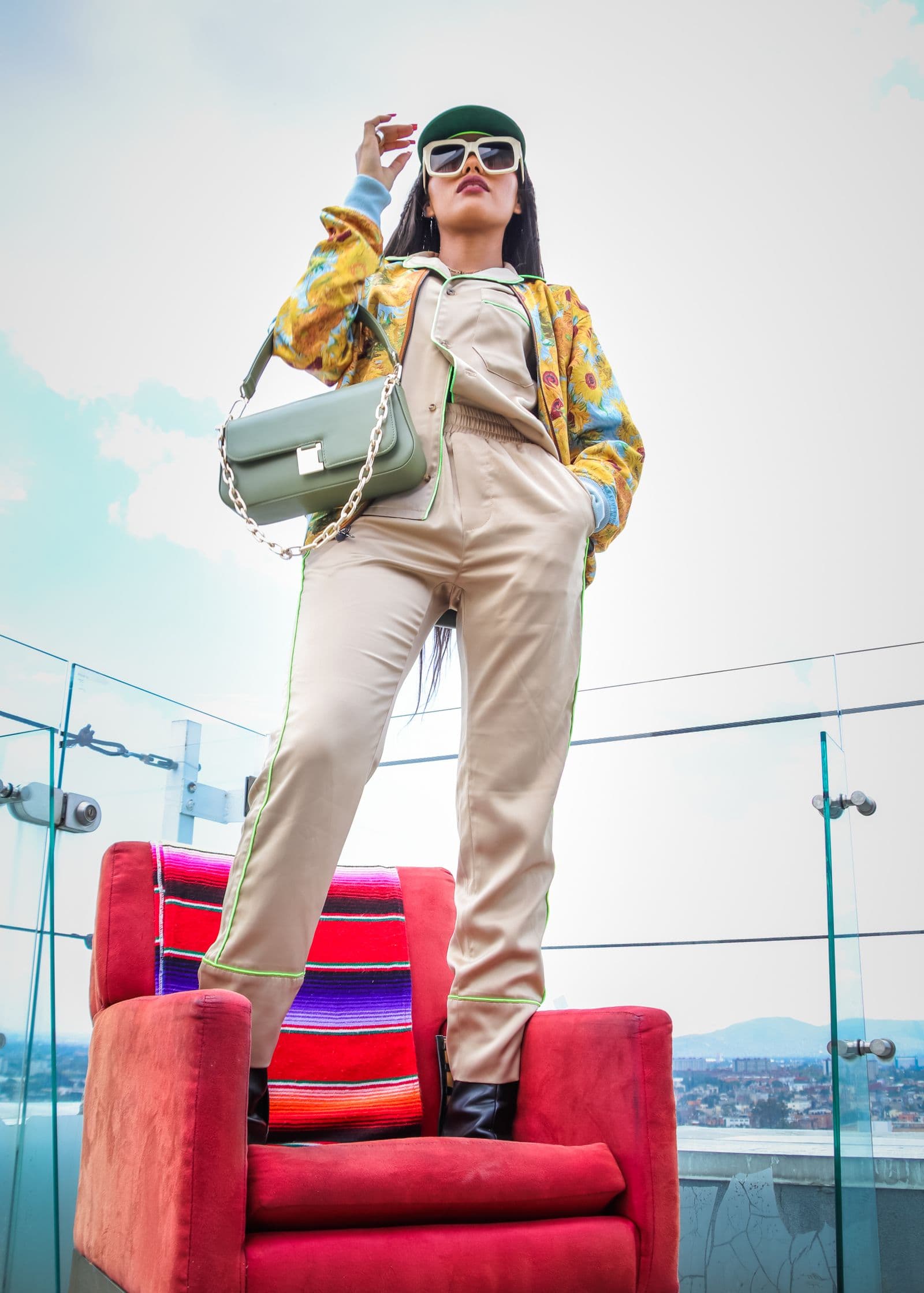 From below, a person in beige clothing and a cap stands on a red armchair on a rooftop under a pale sky.