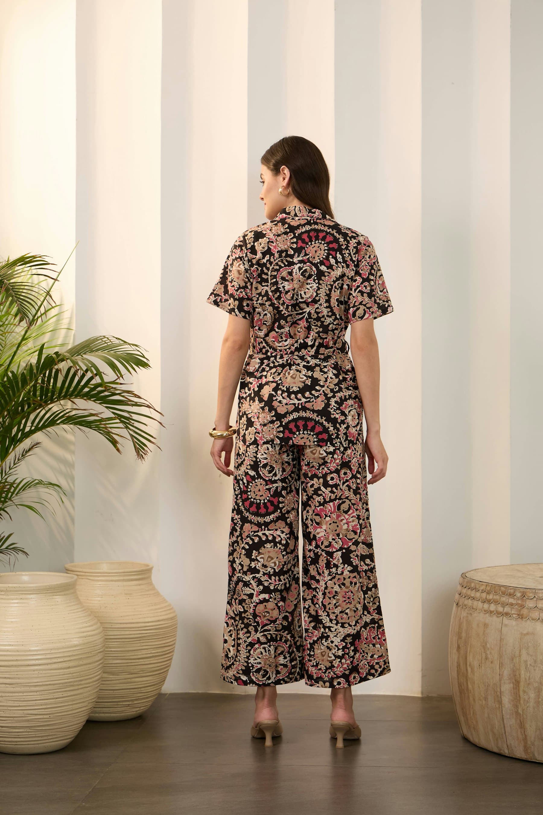 A woman seen from behind wears a floral top with a collar and wide trousers, in a room with a pale wall.