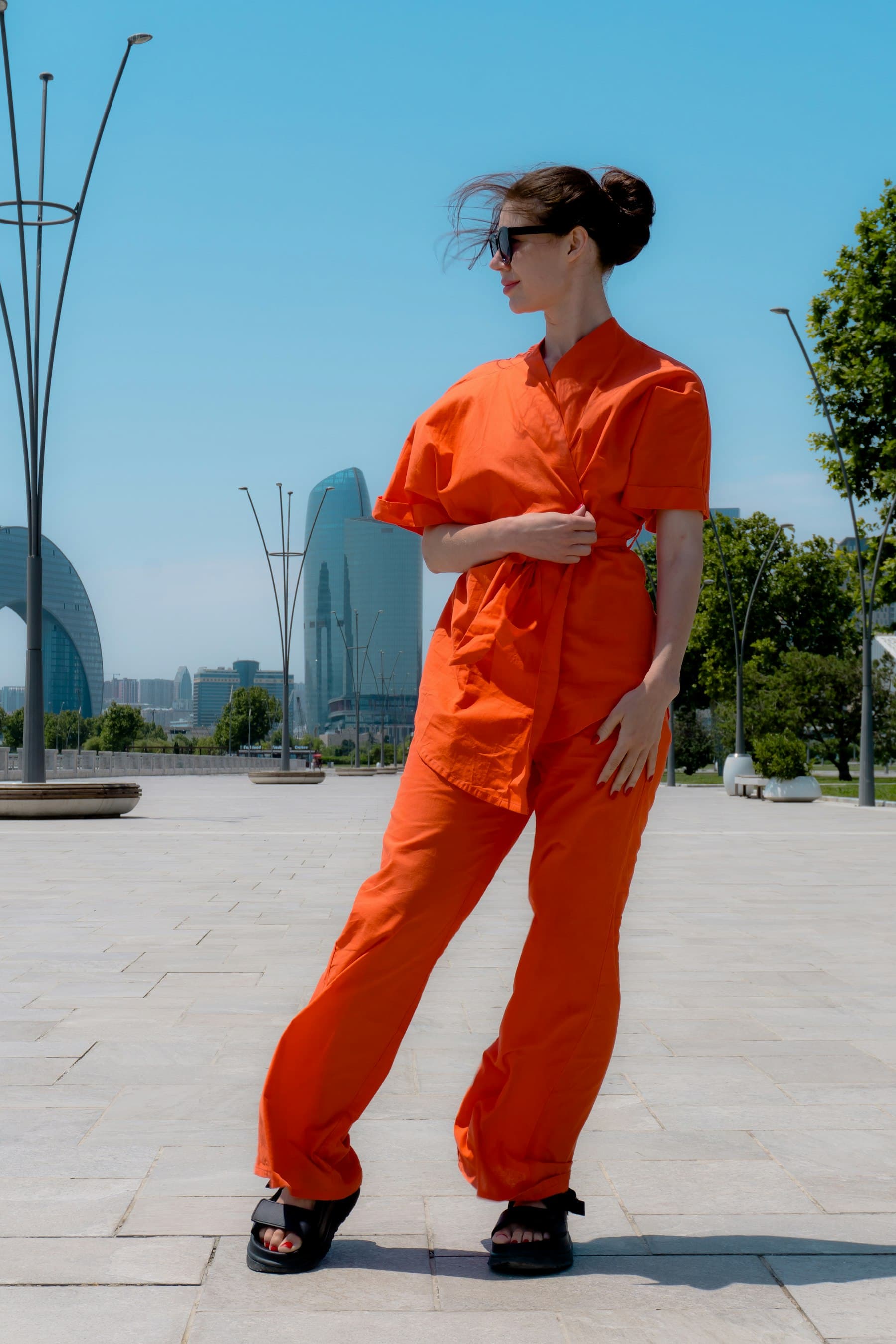 A woman in a bright orange wrap top and wide trousers, wearing dark sunglasses, stands in a plaza with glass towers, trees, and blue sky.