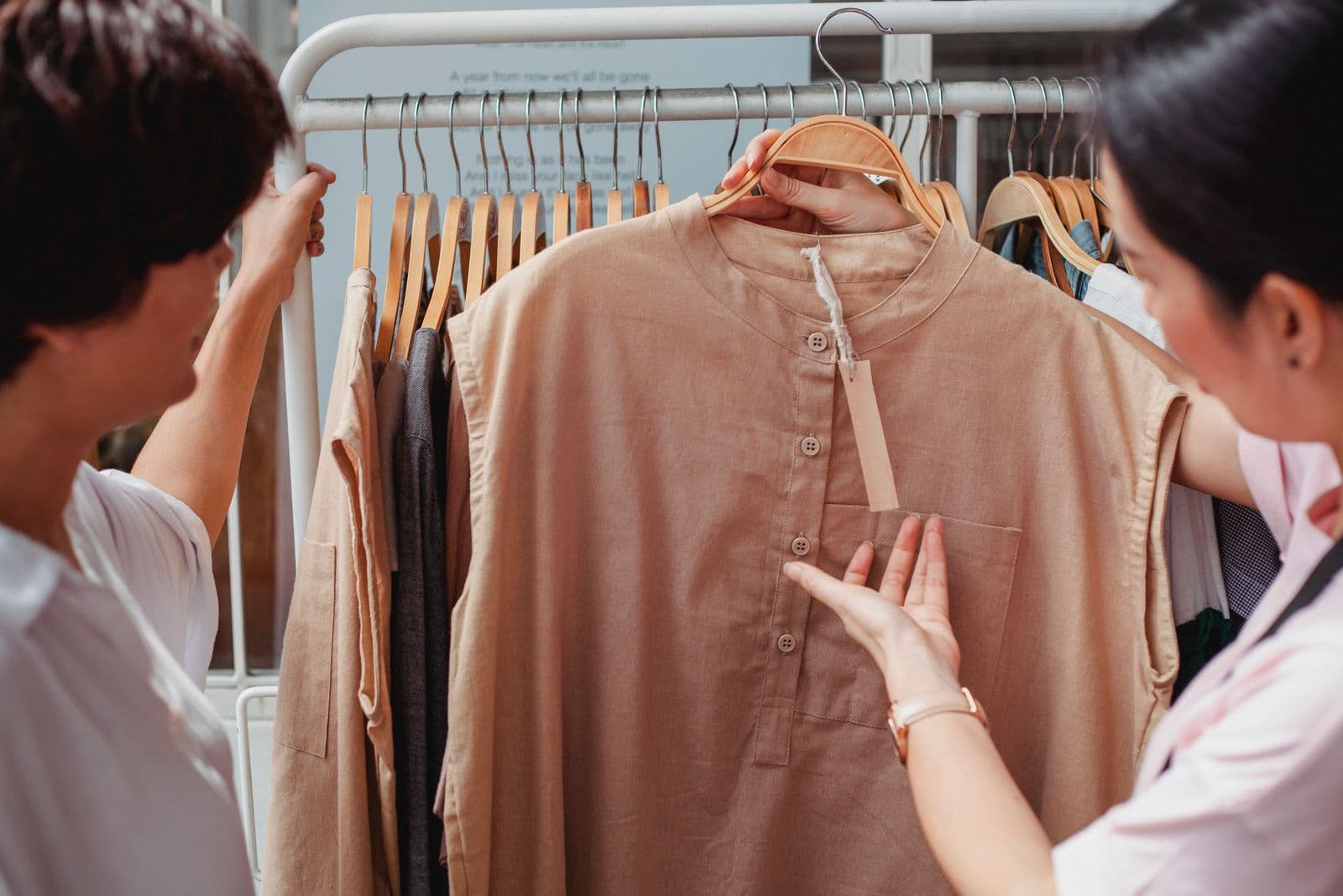 Two people at a clothing rack look over a beige sleeveless tunic with a band collar.
